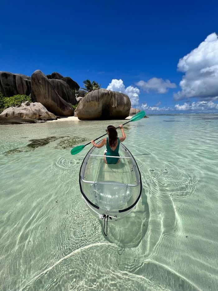 Woman paddling a transparent kayak in clear tropical waters near rocky shore.